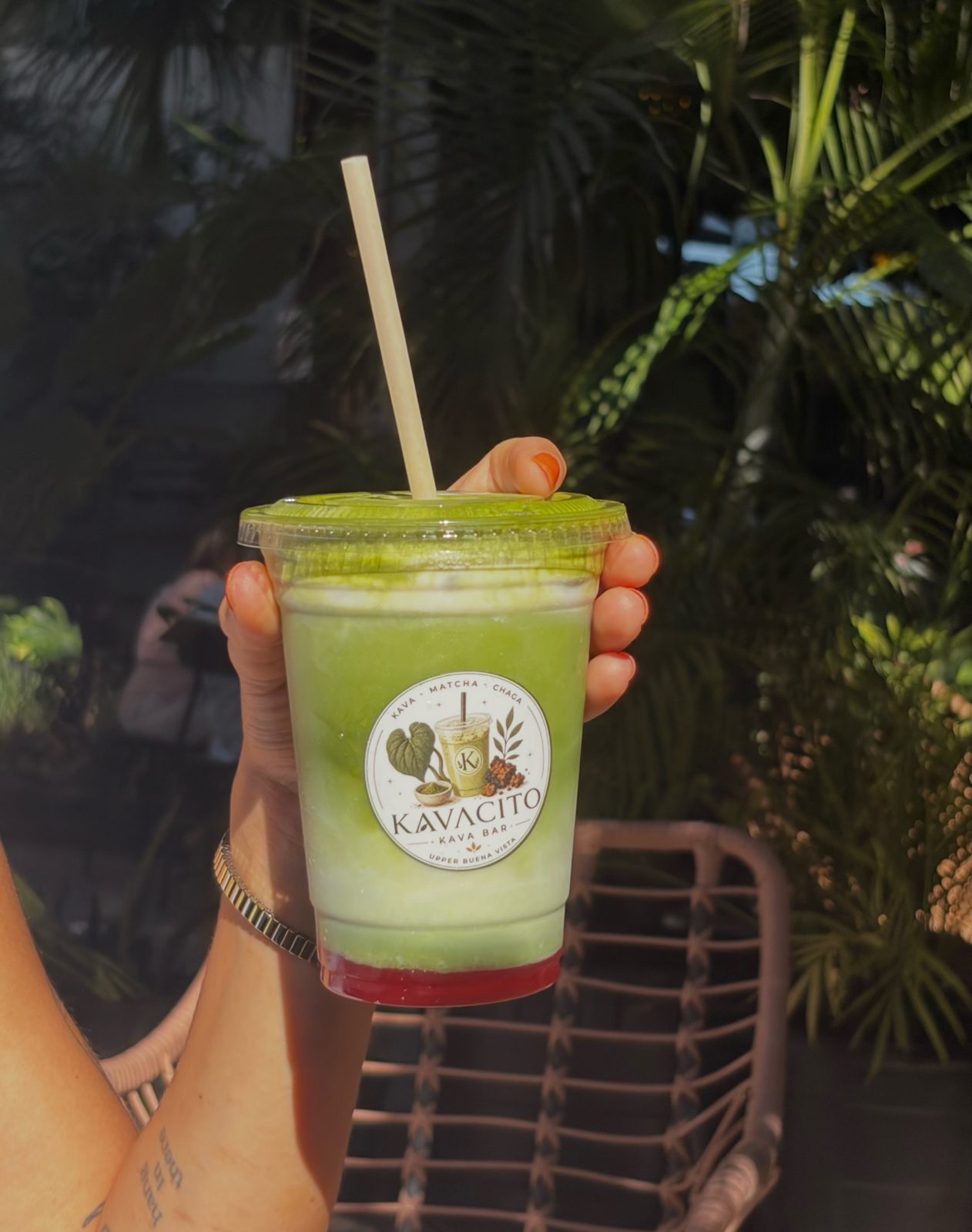 A hand holding a Kavacito matcha beside a rattan chair under the palms