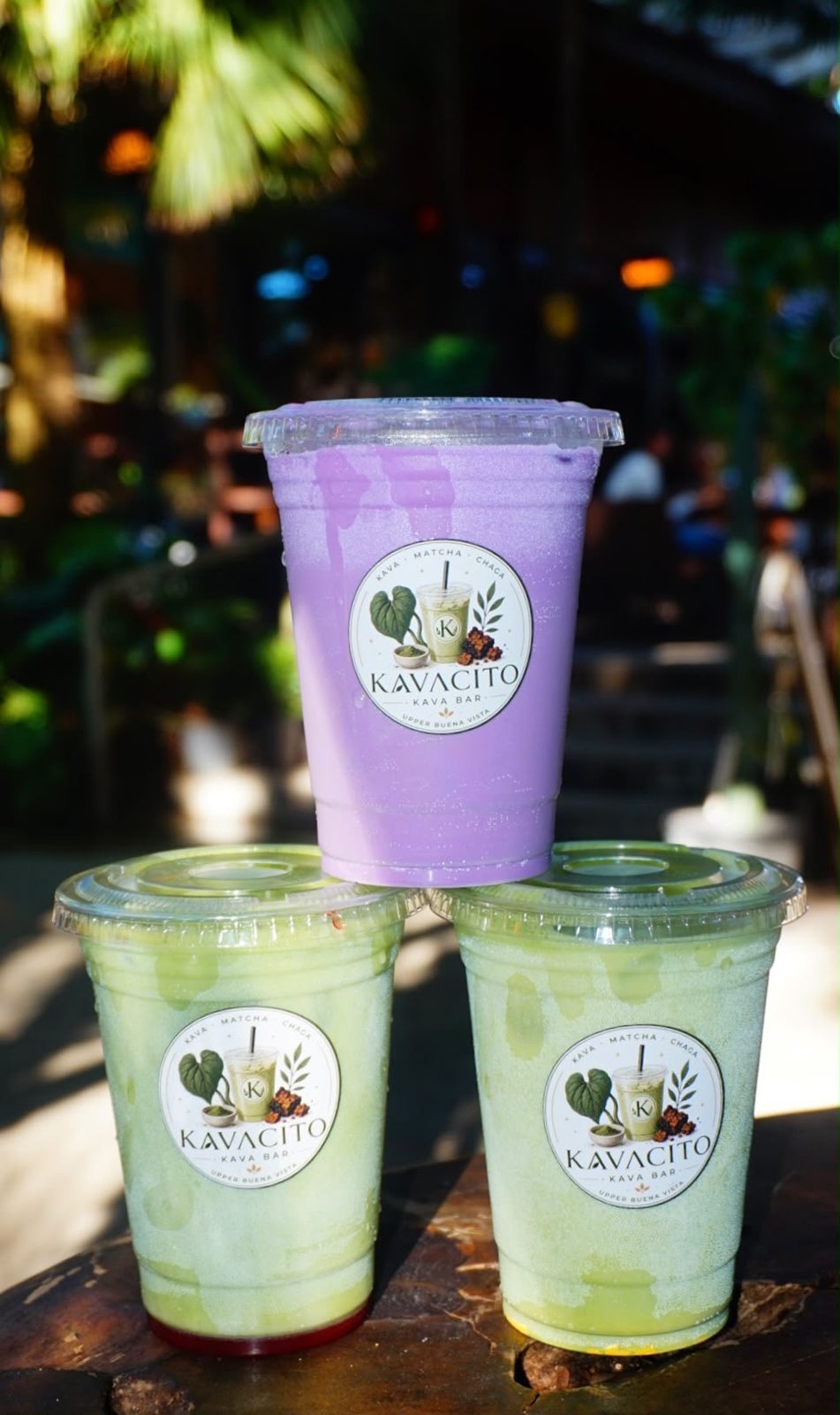 Three Kavacito cups, ube and matcha, stacked on a table with string lights behind